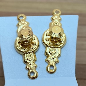 May include: Two gold-toned metal door knobs with ornate designs. Each knob has a round, raised center and a decorative, scroll-like base.