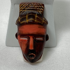 May include: A small, hand-painted mask figurine with a stylized face. The mask features a dark brown face with a red forehead and a woven brown and yellow headdress. The mouth is open, and the eyes are white.