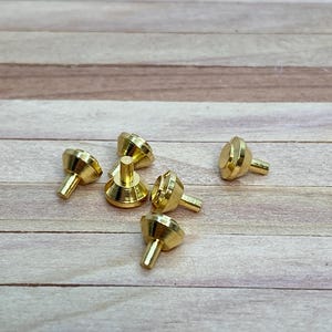 May include: A collection of six small, gold-colored metal objects with a conical shape and a short, cylindrical stem. The objects are arranged on a light brown wooden surface. The items appear to be decorative or functional hardware.