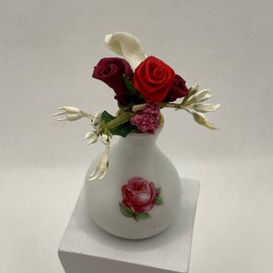May include: A small white ceramic vase with a red rose design, holding a miniature floral arrangement. The arrangement includes red and white fabric flowers, and silver-colored accents. The vase is displayed on a white pedestal.