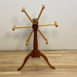May include: Wooden yarn winder with six arms and a tripod base. The winder is made of light brown wood and has a dark brown center.