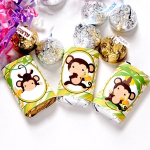 May include: Three chocolate bars wrapped in foil with monkey-themed paper wrappers. The wrappers are green, yellow, and brown with a monkey holding a banana on each. The chocolate bars are sitting on a white surface.