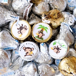 May include: A bowl filled with silver and gold foil-wrapped chocolate kisses, decorated with round stickers featuring cartoon monkeys and jungle-themed designs. The stickers have text that says "Happy Birthday" and "Kisses".