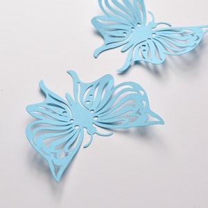 May include: Two light blue paper butterflies with intricate cut-out designs. The butterflies are on a white background.