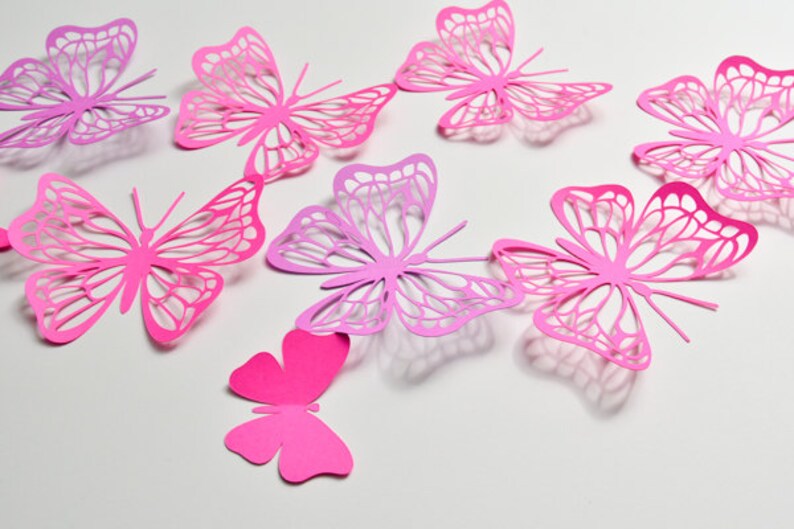May include: A collection of eight pink and purple paper butterflies with intricate cut-out designs. The butterflies are arranged in a random pattern on a white background.