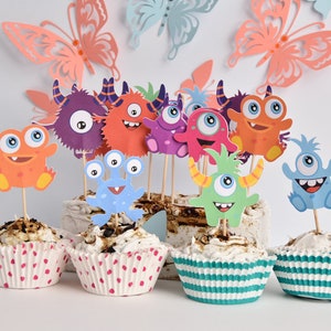 May include: A set of cupcakes decorated with colorful monster-themed toppers. The cupcakes are in pink and green polka dot and striped paper liners. The toppers are made of paper and feature different monster faces with big eyes and smiles.