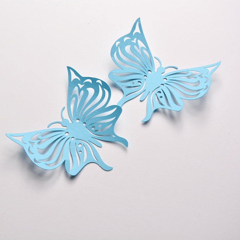 May include: Two delicate blue paper butterflies with intricate cut-out designs. The butterflies are arranged on a white background.