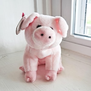 Könnte beinhalten: Ein kleines, hellrosa Plüschschwein mit weicher, flauschiger Textur. Das Schwein hat zwei schwarze Augen, eine Schnauze mit zwei Nasenlöchern und Schlappohren. Am linken Ohr ist ein herzförmiges Etikett mit einem roten Band befestigt.