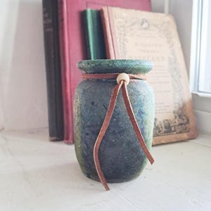 May include: A small, cylindrical ceramic jar with a mottled dark green and blue finish. The jar features a brown leather cord tied around its neck, secured with a single light-colored wooden bead, with two loose ends hanging down the front.