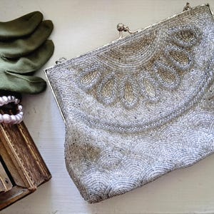 May include: A silver beaded clutch handbag featuring a metallic frame with a kiss-lock closure. The exterior is decorated with intricate silver and gold beadwork arranged in a fan-like pattern at the top and a scalloped design along the bottom.