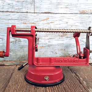 May include: A red metal apple peeler and corer with a wooden handle on the crank. The device features a horizontal threaded rod, a three-pronged metal fork, and a suction cup base. The words BACK TO BASICS are embossed on the side of the frame.