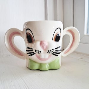 May include: A cream-colored ceramic mug shaped like a rabbit face with two large ear-shaped handles. The rabbit features black eyes, a pink nose, black whiskers, two front teeth, and a light green bow tie at the base of the mug.