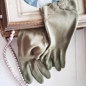 May include: A pair of sage green gloves featuring a scalloped cuff edge with decorative stitching and a small, clear circular button detail at the wrist. The gloves have a smooth, matte finish and are positioned near a string of light pink pearls.