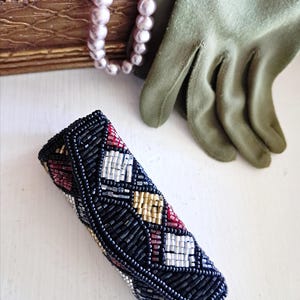 May include: A rectangular beaded case featuring a geometric pattern of black, white, red, and gold glass beads. The case is positioned near a pair of olive green gloves, a strand of light pink pearls, and a textured gold-toned box.
