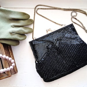 May include: A black beaded handbag with a gold-tone metal frame and a matching gold-tone chain strap. The bag features a geometric pattern of black glass beads. Accompanying the bag are olive green gloves and a strand of pale pink pearls.