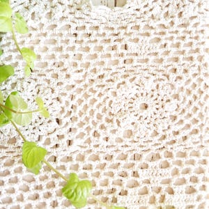 May include: A cream-colored crocheted textile featuring an intricate, repeating geometric pattern with openwork stitches. The fabric displays a circular motif centered within a grid-like structure, showcasing detailed yarn work and a textured surface.