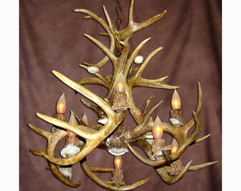 May include: A rustic antler chandelier with multiple candle-style lights. The chandelier is made of natural-colored antlers and has a warm, inviting glow. The fixture is suspended from a chain and is suitable for a cabin or lodge.