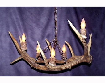 Può includere: Un lampadario rustico a corna di cervo con sei luci a candela finte. Il lampadario è sospeso da una catena e presenta corna marroni e beige. Le luci delle candele hanno un bagliore giallo caldo, creando un'atmosfera accogliente.