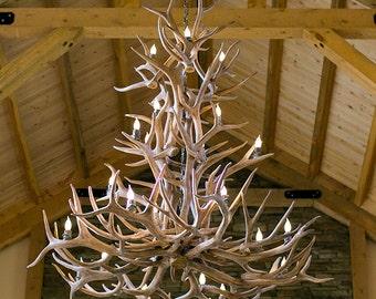 Può includere: Un grande lampadario a corna con più luci a forma di candela. Il lampadario è fatto di corna colorate naturalmente e pende da una catena. Le luci forniscono un'illuminazione calda. L'apparecchio è sospeso a un soffitto di legno.