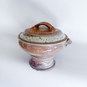 Puede incluir: Un plato de servir de cerámica con tapa y asa. El plato es marrón y blanco con motas y tiene una base de pedestal.