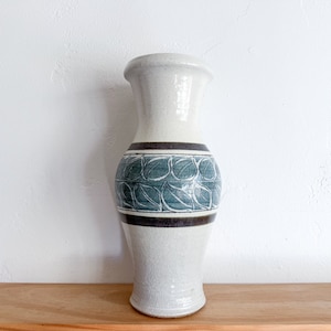 Op de afbeelding: Een witte keramische vaas met een blauw en bruin bladmotief. De vaas heeft een rond lichaam en een smalle hals.