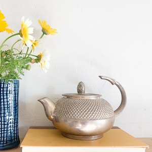 May include: A bouquet of yellow and white daisies in a blue and white patterned ceramic vase, next to a silver teapot with a textured surface on a yellow surface.