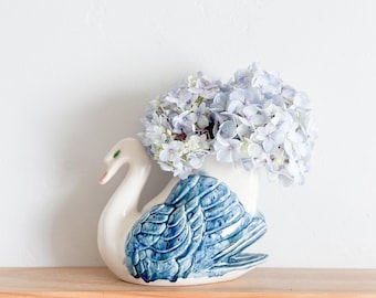 May include: A white ceramic swan vase with blue and white painted details, holding a bouquet of light blue hydrangeas.