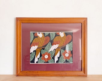 May include: A framed artwork depicting two stylised birds with brown, orange and white feathers perched on branches. The birds are surrounded by green leaves and red flowers against a black background.