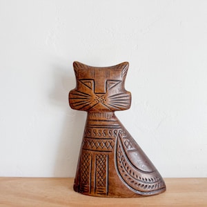 May include: A wooden cat figurine with a brown and black geometric pattern. The cat has a stylized face with a long nose and large eyes.