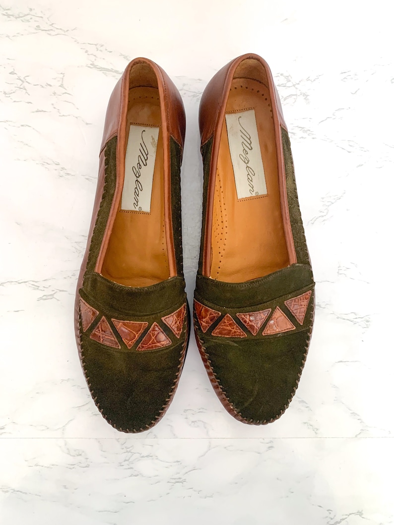 May include: A pair of brown leather loafers with green suede uppers and brown leather accents. The loafers have a pointed toe and a low heel. The brand name "Morgan" is printed on the inside of each shoe.
