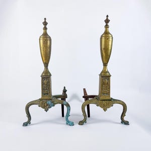 May include: A pair of antique brass fireplace andirons with ornate details and a decorative urn design. The andirons have a distressed finish and are in good condition.