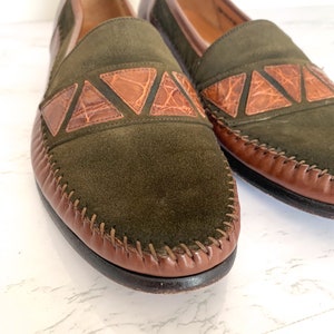 May include: A pair of green suede loafers with brown leather trim and a crocodile pattern detail. The loafers have a stitched edge and a brown leather sole.