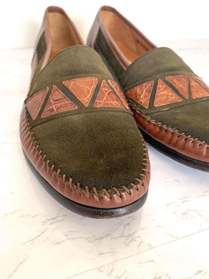 May include: A pair of green suede loafers with brown leather trim and a crocodile pattern detail. The loafers have a stitched edge and a brown leather sole.
