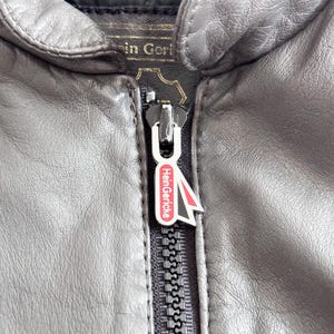Op de afbeelding: Close-up van een zwarte rits met een zilveren rits en een rood en wit logo met de tekst "HeinGericke".