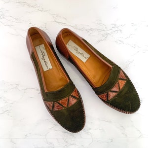 May include: A pair of brown leather loafers with green suede uppers and brown leather accents. The loafers have a pointed toe and a low heel. The brand name "Meg-Lane" is printed on the insole.