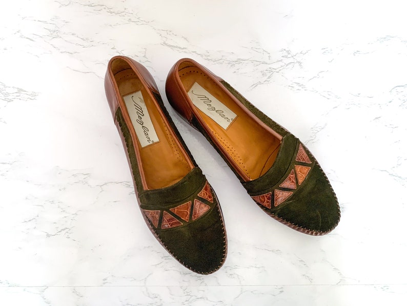 May include: A pair of brown leather loafers with green suede uppers and brown leather accents. The loafers have a pointed toe and a low heel. The brand name "Meg-Lane" is printed on the insole.