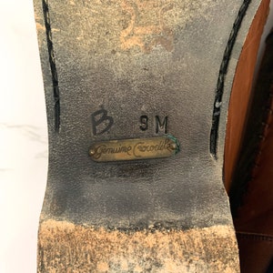 May include: Close-up of the sole of a brown leather shoe with a black stitching detail. The shoe has a label that reads "Genuine Crocodile" and the size "9M" is written on the sole.