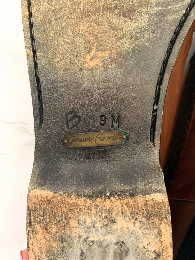 May include: Close-up of the sole of a brown leather shoe with a black stitching detail. The shoe has a label that reads "Genuine Crocodile" and the size "9M" is written on the sole.
