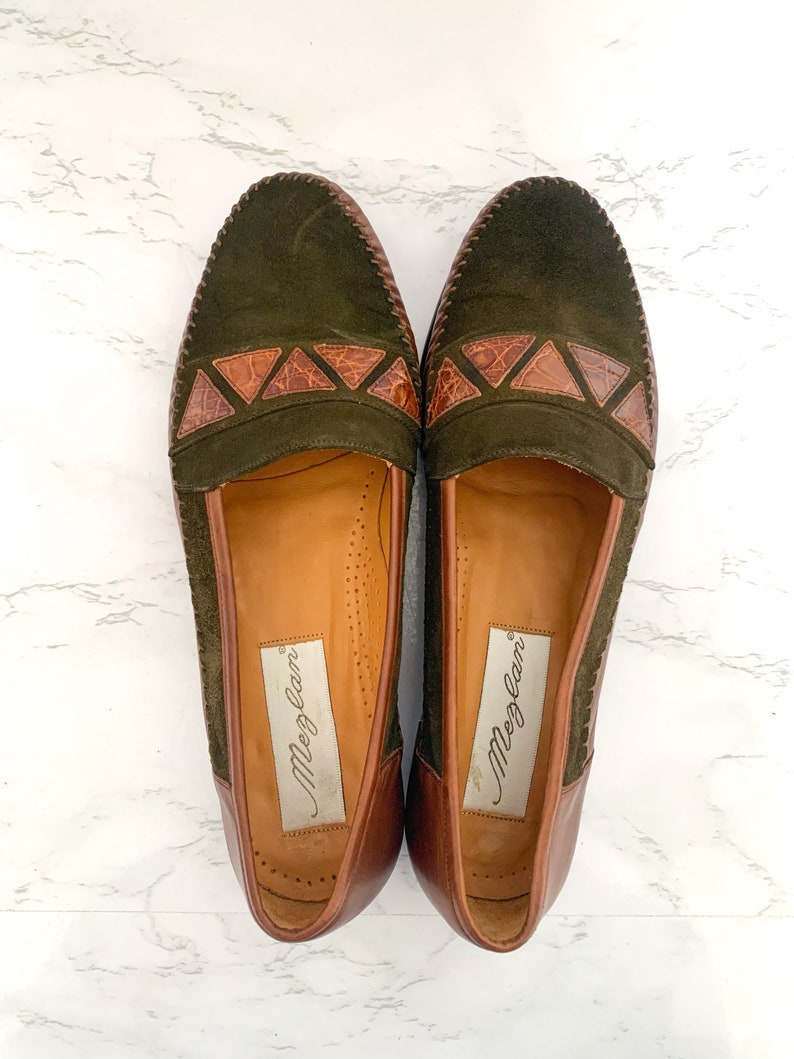 May include: A pair of green suede loafers with brown leather accents and the brand name "Meglan" on the inside.