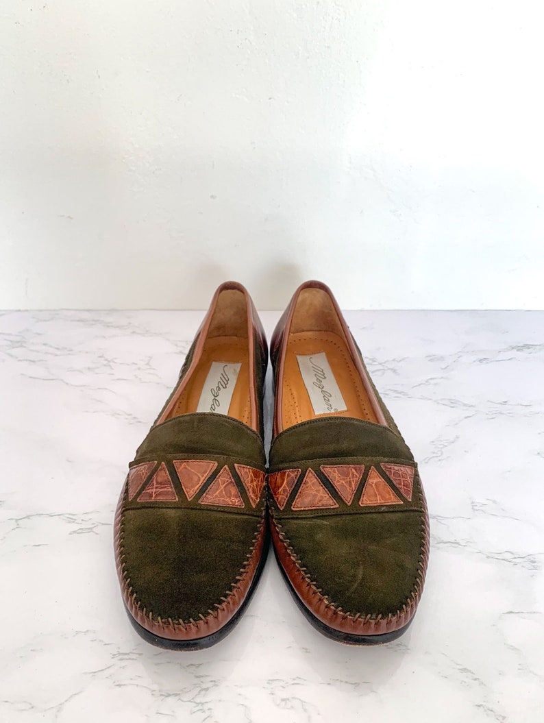 May include: A pair of green suede loafers with brown leather trim and a crocodile-embossed detail. The loafers are by the brand "May & Law".