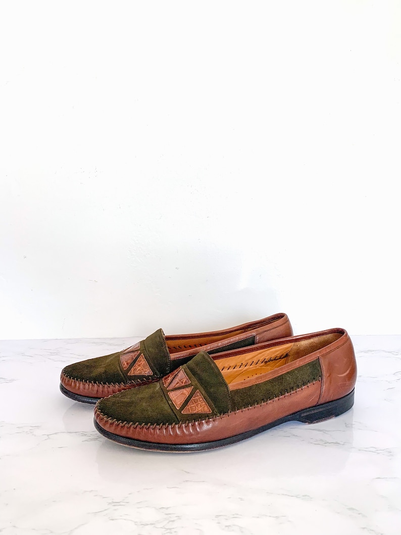 May include: A pair of brown leather loafers with green suede uppers and decorative brown leather accents. The loafers have a stitched edge and a brown leather sole.