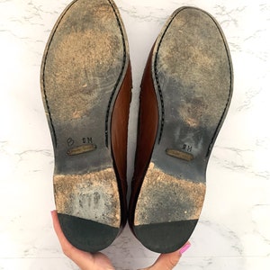 May include: A pair of brown leather shoes with black soles. The shoes are size 9M.