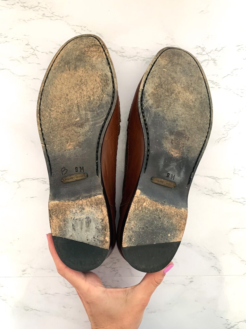 May include: A pair of brown leather shoes with black soles. The shoes are size 9M.