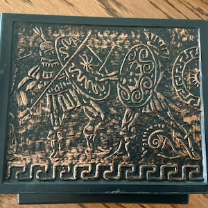 May include: A black box with a bronze-colored relief depicting ancient warriors with shields and spears. The box has a decorative border and a dark finish, suggesting a historical or antique theme.