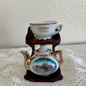 May include: A miniature ceramic teapot and teacup set. The teapot features a gold handle and spout, with a Boston, Massachusetts scene. The teacup is white with gold trim and the words "BOSTON MASS." on the side. The set is displayed on a small wooden shelf.