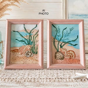 May include: Two framed cross-stitch artworks featuring coastal scenes with green seaweed, tan sand, and various seashells. Each piece is encased in a light pink wooden frame. The text AI PHOTO is visible in the centre between the two frames.