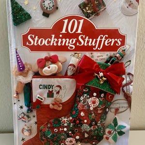 May include: A book cover titled "101 Stocking Stuffers" with a red banner. The cover features a Christmas stocking filled with ornaments, a teddy bear, and a Santa Claus figure. The stocking is red with green and red embellishments.