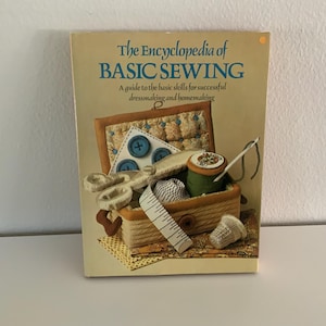 May include: A vintage book titled "The Encyclopedia of BASIC SEWING" with a guide to basic skills for dressmaking and homemaking. The cover features a sewing box with spools of thread, buttons, and a thimble. The book is tan with blue text.