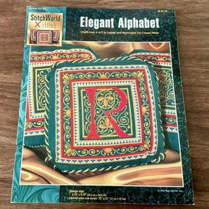 May include: A cross-stitch pattern book titled "Elegant Alphabet" featuring a pillow design with the letter "R". The pillow design is green, gold, and red, with a finished size of 13 inches by 13 inches.
