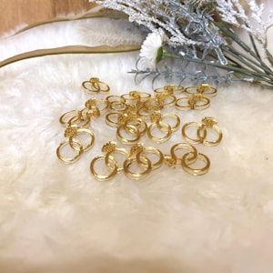 May include: A collection of small, gold-colored rings, some with a diamond-shaped embellishment, are scattered on a soft, white, textured surface. The rings are arranged in pairs and singles. Decorative greenery with white flowers is in the background.